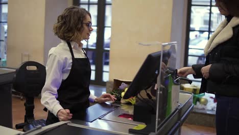 Smiling-woman-customer-paying-with-her-EC-card-at-supermarket-checkout-and-taking-her-brown-paper-bag-with-products-from-the-counter.-Female-cashier-standing-in-black-apron
