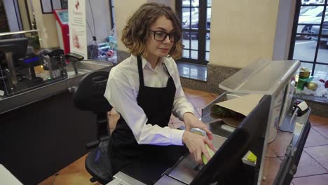 Cheerful-slender-saleswoman-in-white-shirt-and-black-apron-scanning-product,-fruits-at-checkout-counter-in-bright-supermarket-and-putting-it-into-brown-paper-bag.-High-angle-footage