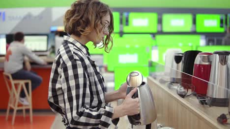 Side-view-of-a-young-cheerful-positive-attractive-woman-in-plaid-shirt-choosing-electronic-kettle-in-household-appliances-store,-standing-in-a-row,-holding-and-examines-silver-kettle