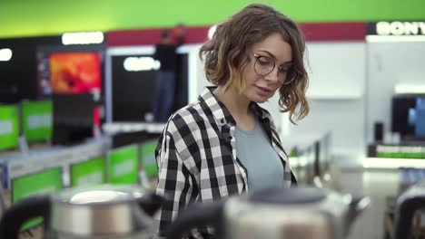 Young-cheerful-positive-woman-in-plaid-shirt-and-glasses-choosing-electronic-kettle-in-household-appliances-store,-walking-in-a-row,-holding-and-examines-silver-kettle