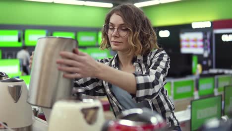 Young-cheerful-positive-woman-in-plaid-shirt-and-glasses-choosing-electronic-kettle-in-household-appliances-store,-taking-one-from-the-shelf-and-examines-silver-kettle