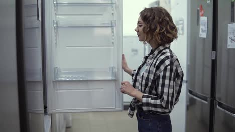 Cheerful-young-girl-selecting-domestic-refrigerator-in-supermarket.-Curly-woman-in-plaid-shirt-deciding-to-buy-a-new-one-for-home