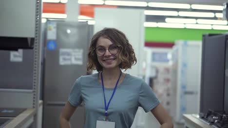 Positive-female-seller-or-shop-assistant-portrait-in-supermarket-store.-Woman-in-blue-shirt-and-empty-badge-looking-at-the-camera-and-smiling.-Household-appliances-on-the-background
