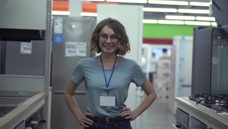 Positive-female-seller-or-shop-assistant-portrait-in-supermarket-store.-Woman-in-blue-shirt-and-empty-badge-looking-at-the-camera-and-smiling.-Household-appliances-on-the-background.-Close-up-camera