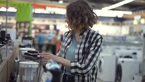 In-the-appliances-store,-a-brunette-curly-woman-in-a-plaid-shirt-chooses-a-blender-for-shopping-by-viewing-and-holding-the-device-top-in-her-hands.-Side-view