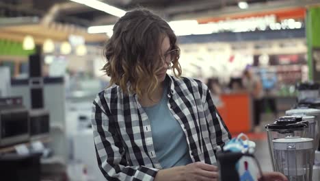 In-the-appliances-store,-a-brunette-curly-woman-in-a-plaid-shirt-chooses-a-blender-for-shopping-by-viewing-and-holding-the-device-top-in-her-hands.-Slow-motion