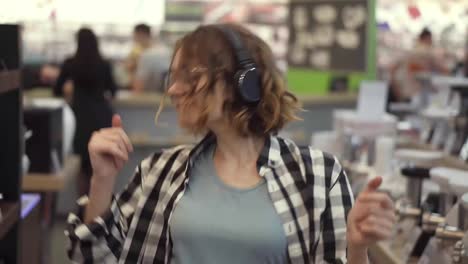 Portrait-of-cute-caucasian-pretty-young-woman-with-short-curly-hair,-plaid-shirt-and-in-headphones-dancing-and-listening-music-in-the-middle-of-the-supermarket-with-household-stuff.-Slow-motion