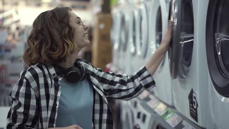 A-young-positive-woman-in-a-plaid-shirt-choosing-washing-machine-in-the-shop-of-household-appliances.-Opens-the-door-and-look-inside.-Side-view