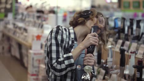 Portrait-of-cute-caucasian-pretty-young-woman-with-short-curly-hair,-plaid-shirt-and-in-headphones-dancing-and-pretending-she's-singing-in-the-middle-of-the-supermarket-with-household-using-blender-stick-as-microphone