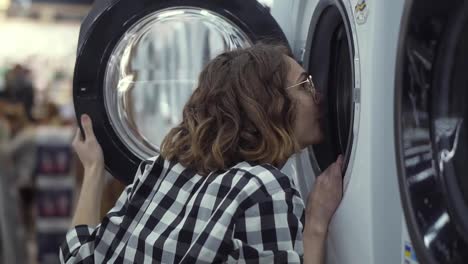 A-young-positive-woman-in-a-plaid-shirt-choosing-washing-machine-in-the-shop-of-household-appliances.-Look-inside-to-the-open-door-and-smiling.-Side-view.-Slow-motion