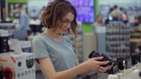 Young-woman-in-glasses-shopping-for-a-new-photocamera-in-the-electronics-store.-Trying-to-decide-on-the-best-model.-Have-Doubts.-Checking-one-camera-in-hands