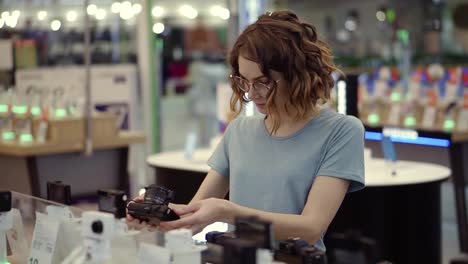 Young-curly-woman-in-glasses-shopping-for-a-new-photocamera-in-the-electronics-store.-Trying-to-decide-on-the-best-model.-Have-Doubts.-Checking-one-camera-in-hands