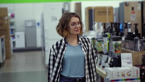 Tracking-front-view-footage-of-a-girl-in-a-plaid-shirt-and-short-curly-hair-walking-by-supermarket-in-household-appliences-looking-for-something.-Slow-motion