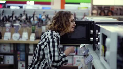 Side-view-of-a-young-curly-haired-woman-in-plaid-shirt-chooses-a-microwave-oven-in-a-consumer-electronics-supermarket.-Opens-the-door-and-looking-inside-the-oven