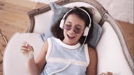 Stylish-hipster-girl-in-her-20's-wearing-headphones,-glasses-and-punk-leather-biker-gloves,-lying-on-the-couch,-singing-and-dancing-to-the-music