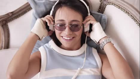 Attractive-cheerful-girl-with-headphones-and-glasses-smiling-and-dancing-to-the-music-while-lying-on-the-sofa