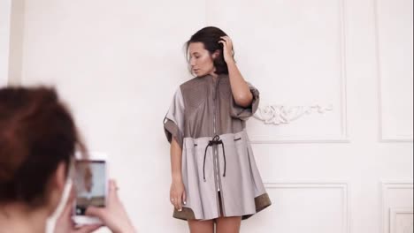 Unrecognizable-girl-with-hair-pulled-in-a-bun,-taking-picture-with-a-smartphone-of-a-young-stylish-girl,-wearing-elegant-leather-dress,-standing-on-white-wall-background