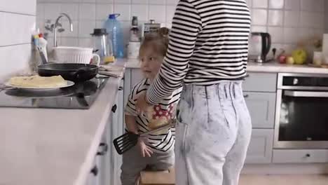 Young-stylish-mother-helps-daughter-turn-the-pancake-with-a-shovel-and-having-fun-while-cooking-together-in-kitchen-at-home-with-two-children
