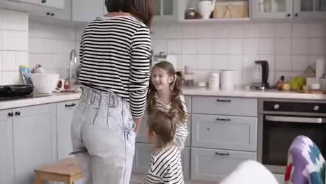 Funny,-young-and-cheerful-mother-is-playing-with-daughters.-Imitating-horse-riding-using-shovels-in-the-kitchen-while-cooking
