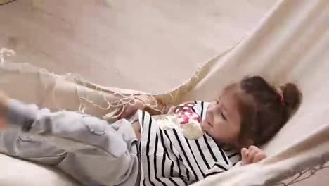 Happy-little-girl-having-fun-indoors,-lying-in-hammock,-and-waving-her-legs.-Smiling-child