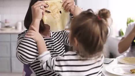 Funny-mother-is-playing-with-her-little-daughter-making-comical-mask-with-pancake-on-her-face.-Family-breakfast