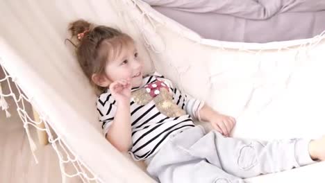 Little-smiling-girl-chilling-and-swinging-on-a-white-hammock.-Indoors
