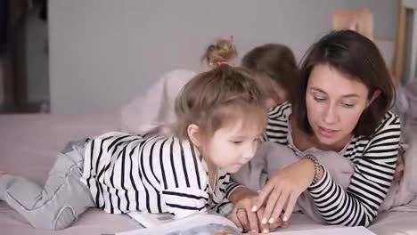 Young-mother-is-reading-a-tale-for-her-lovely-little-daughters-lying-on-a-bed.-Little-one-is-looking-through-the-book.-Close-up