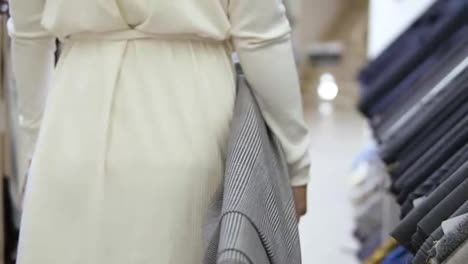 Tracking-footage-of-a-beautiful-woman-in-white,-walking-by-the-cloth-store-with-a-roll-of-light-textile-in-the-right-hand.-Close-up-shot-from-back-side