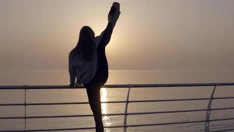 Slim-woman-with-long-legs-doing-stretching-on-sunrise-by-the-seaside,-perfect-body.-Stretches-her-leg-above-head.-Backside-view
