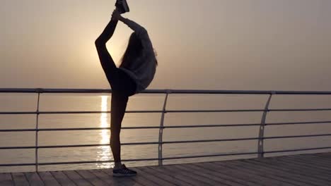 Long-haired-girl-stretches-her-leg-with-a-view-of-the-sea-ocean.-Dawn-Sunrise.-Sportswear,-leggings,-hoodies.-Dusk