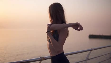 Close-up-of-a-beautiful-young-girl-in-a-sports-bra-stretches-her-arm-muscles.-Long-haired-blonde.-The-sun-is-shining-above-the-sea-ocean.-Sport-life