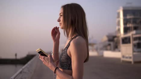 A-bright,-slender-girl-stands-in-the-morning-near-the-sea,-puts-on-headphones,-a-mobile-phone-in-her-hands.-Preparing-for-training.-In-a-sports-bra.-Overview-footage