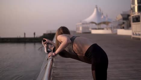 Thin-and-fit-young-female-in-sports-clothes-stretching-leg-muscles-on-a-sea-area.-Wearing-black-leggings-and-grey-bra.-Leans-on-a-crossbar.-Side-view