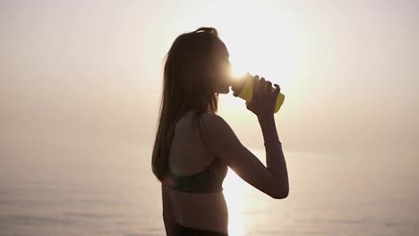 Young-long-haired-woman-runner-having-a-break,-drinking-water-against-clear-blue-sky-and-sun-background,-jogging.-Sports-bra