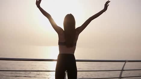 Cheerful,-young-woman-opens-arms-to-sunrise-at-seaside.-Slim-and-graceful,-in-leggings.-Back-Side-view