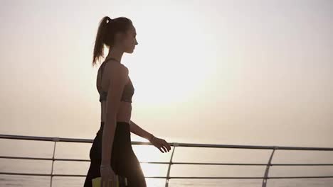Gorgeous-side-view-of-a-pretty-girl-walking-on-the-road-along-the-sea,-the-sun-shines-quietly.-A-girl-in-black-leggings-and-top.-Pony-tail.-Carefree