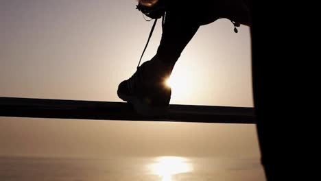 Extremely-close-up.-Silhouette-of-a-slender-girl,-ties-up-shoelaces-on-sneakers-leaning-on-the-crossbar.-Morning-sunrise-on-the-background