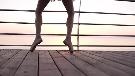Aim-footage-of-a-ballet-dancer's-feet-while-she-practices-pointe-exercises-on-the-wooden-embankment-near-sea.-Close-up-of-woman's-legs-in-pointe-shoes.-Sunrise.-Slow-motion