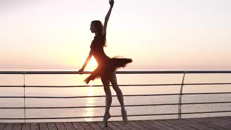 Full-length.-A-ballet-dancer-in-a-black-tutu,-doing-exercises,-leaning-on-a-bar-near-the-sea.-Young-ballet-dancer-stretches-her-legs-in-a-squat.-Morning,-pink-sunrise.-Slow-motion