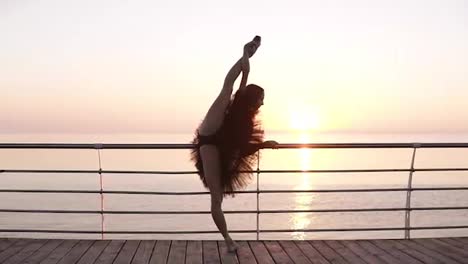A-ballerina-is-standing-near-the-banister-on-the-promenade.-She-is-stretching,-bending-her-legs-in-a-vertical-twine.-Sun-shines.-Beautiful-morning-marine-view