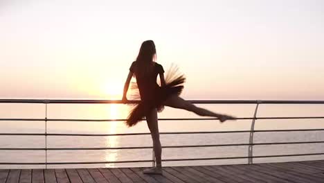 Ballerina-stands-facing-to-the-sea-or-ocean-in-a-black-tutu.-Young-beautiful-woman-practicing-stretching-and-exercises.-Sun-almost-raised.-Backside-view