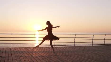 Beautiful-silhouette-of-ballerina-in-ballet-tutu-and-point-on-embankment-above-ocean-or-sea-at-sunrise.-Young-beautiful-long-haired-woman-practicing-and-exercises.-Morning-bosk
