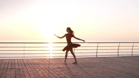 Ballerina-in-black-ballet-tutu-and-point-on-embankment-above-ocean-or-sea-at-sunrise.-Jumping-ballerina,-practicing-classic-exercises.-Slow-motion