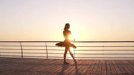 Attractive-ballerina-practices-in-stretching.-Doing-classic-ballet-moves.-Long-haired-young-girl-in-dark-tutu.-Embankment-near-the-sea-or-ocean.-Sun-shines.-Slow-motion