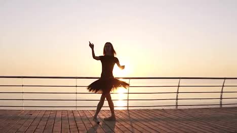 Dancing-ballerina-in-black-ballet-tutu-and-pointe-on-seafront-above-ocean-or-sea.-Morning-bosk.-Young-attractive-woman-practicing,-making-vertical-twine,-lunge-forward-in-the-dance