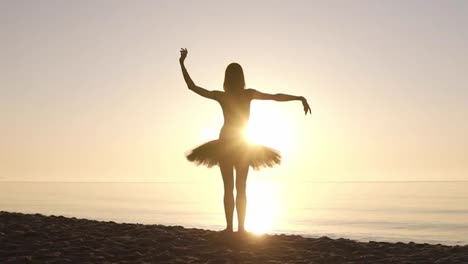 Graceful-young-girl-in-ballet-tutu-standing-on-a-coastline-facing-to-the-sea.-Waving-her-hands.-Doing-ballet-moves.-Sun-shines.-Backside-view