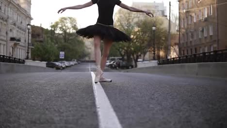 Ballerina-in-black-ballet-tutu-and-point-on-empty-street-in-the-middle-of-the-road.-Summer-morning.-Young-beautiful-woman-practicing-ballet-movements-and-exercises.-Close-up.-No-face