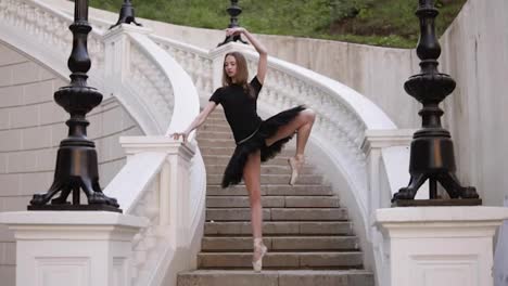 Front-view-of-a-graceful-ballet-movements.-Young,-blonde-haired-ballerina-doing-vertical-twine-and-posing-standing-on-a-beginning-of-street-stairs-in-black-tutu.-Daytime.-Slow-motion