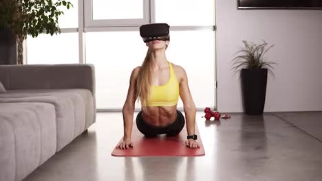 Long-haired-woman-with-muscular-body-wears-VR-mask-during-her-home-work-out-at-living-room.-Girl-in-a-mask-doing-stretching-workout-on-mat-at-home.-Front-view