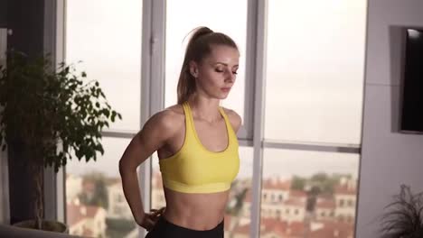 Young-female-athlete-is-warming-up-in-sportswear-at-her-living-room.-She-is-leaning-torso-forward,-stretching-hands-locked-benind-back.-Cropped-footage.-Domestic-workout